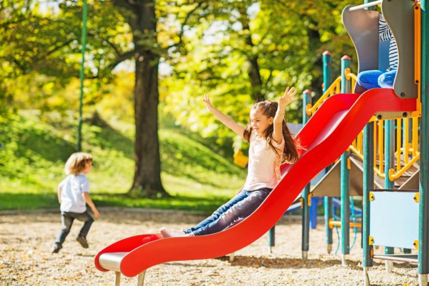 Man sieht mehrere Kinder, die draußen spielen. Ein Mädchen rutscht eine rote Rutsche runter.